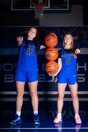 Two basketball players in blue uniforms pose with basketballs on a court under a hoop.