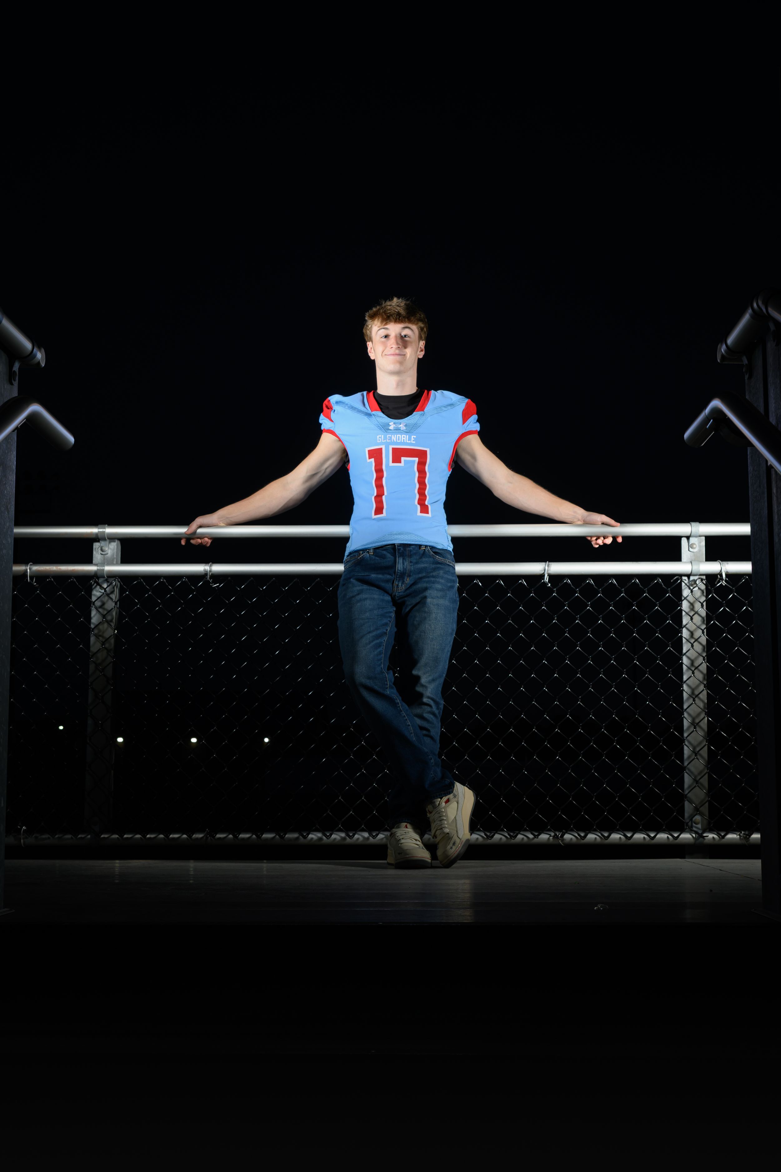 A person in a jersey with number 17 sits on a metal railing against a dark background.