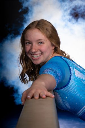 Gymnast in blue sparkly leotard performs on balance beam with dramatic smoke effect in background.