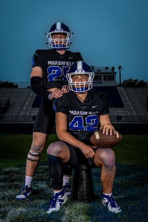 Two football players in matching black jerseys pose on field with stadium bleachers in background at dusk.