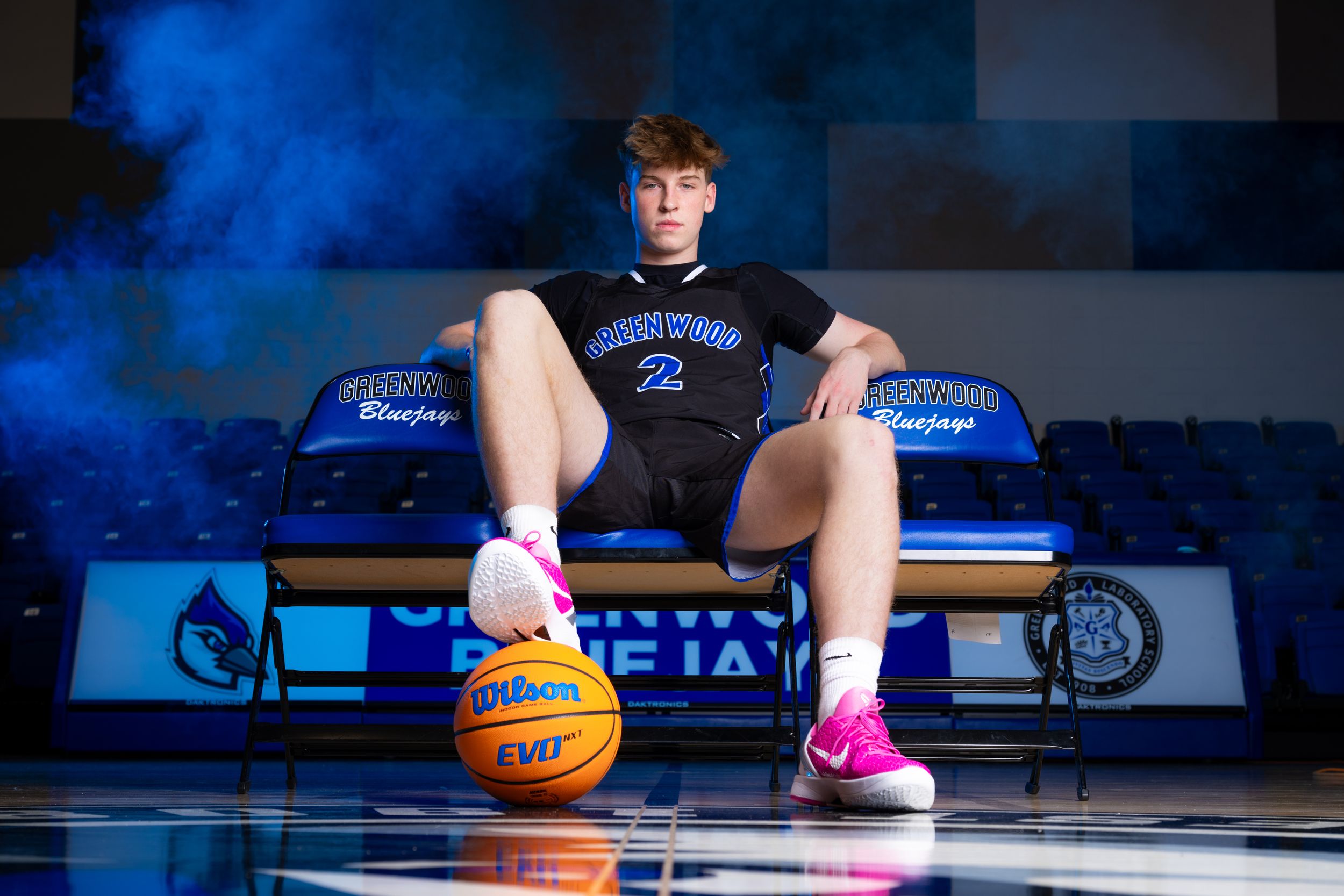 Basketball player sits on bleachers with a ball in dramatic blue lighting on indoor court.