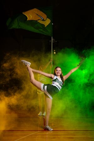 Cheerleader performs high kick pose with colorful smoke effects in green and orange.