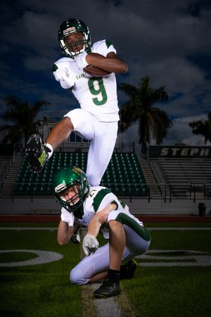 Football players execute dramatic jumping play on field at night with stadium lights.