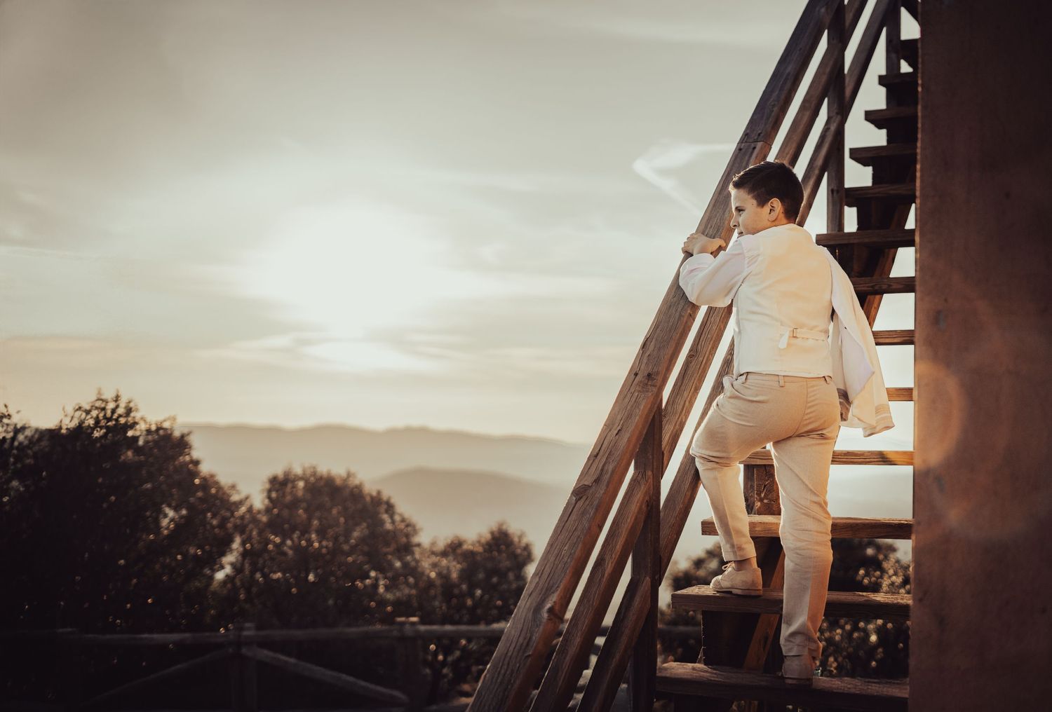 Boy in white communion suit climbing wooden stairs with mountain sunset glowing in background.