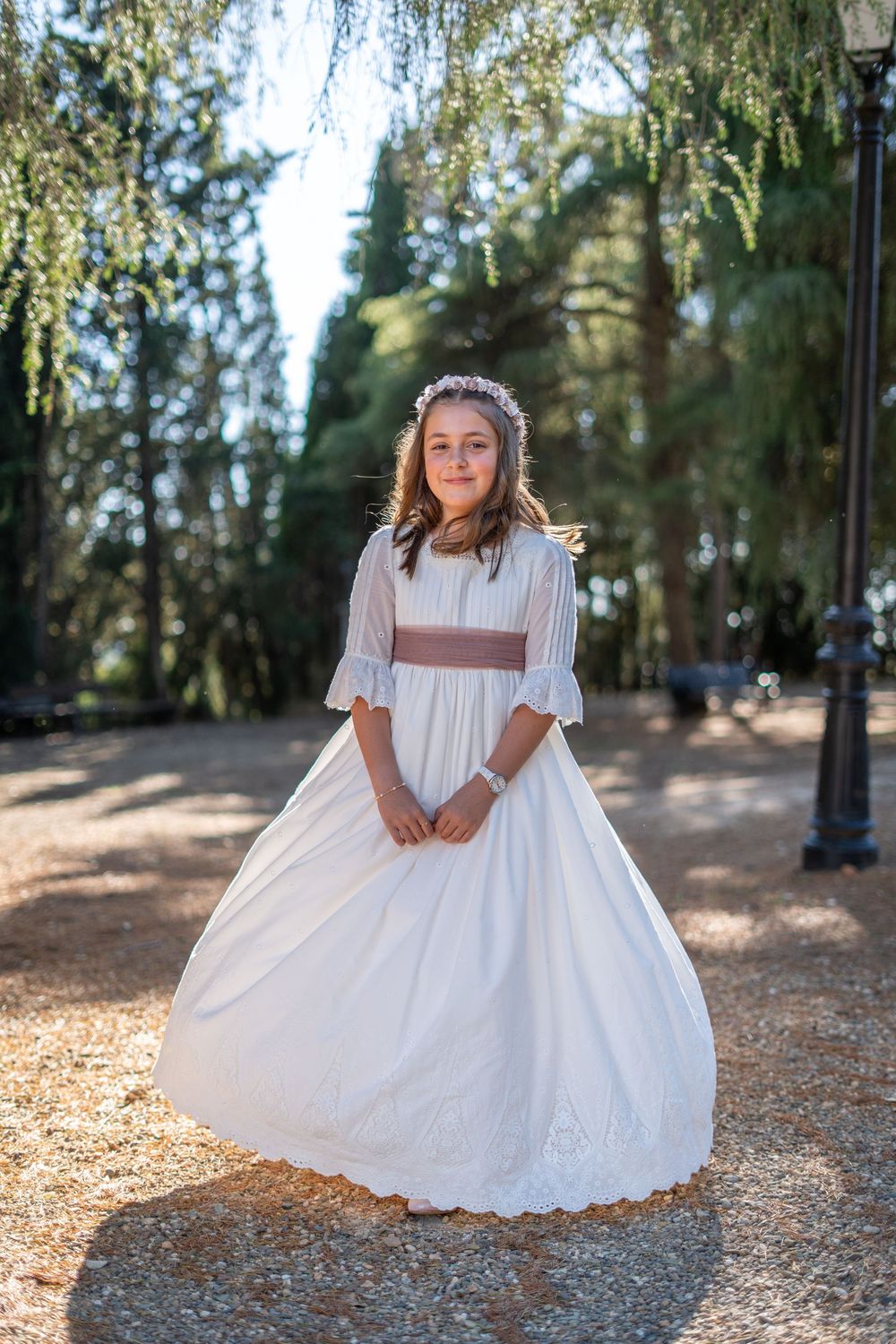 Young girl in white communion dress and floral headband posing in a sunlit park with tall trees.