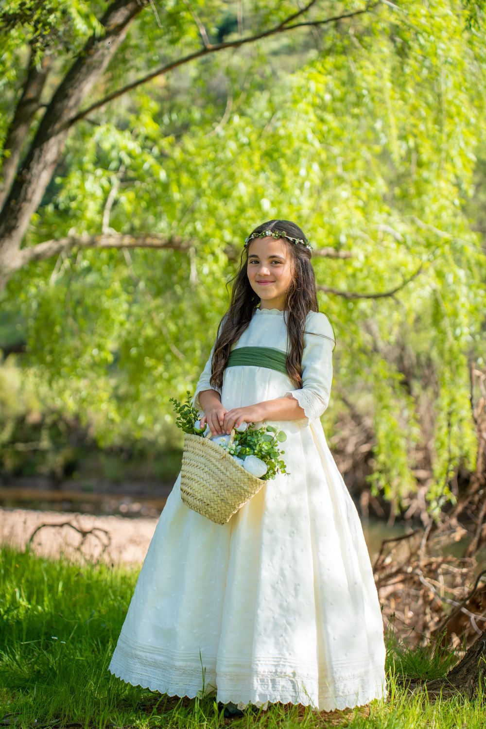 Young girl in white communion dress holding flower basket, wearing floral crown, standing in lush green garden.