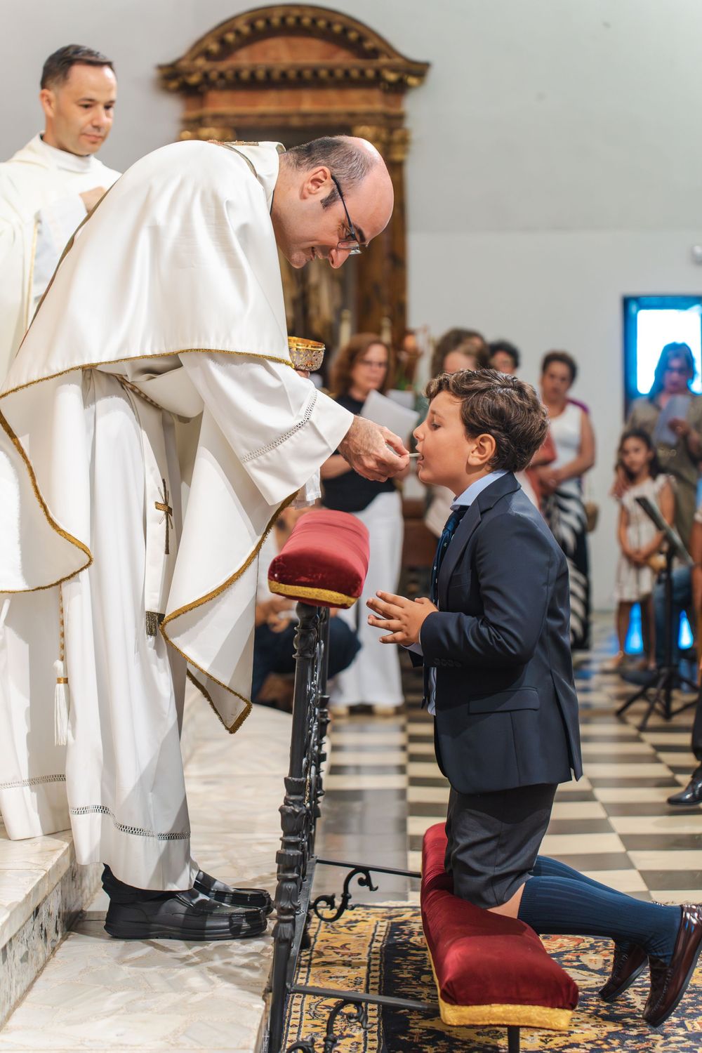 Priest administering First Communion to a young boy kneeling at altar in a Catholic church ceremony.