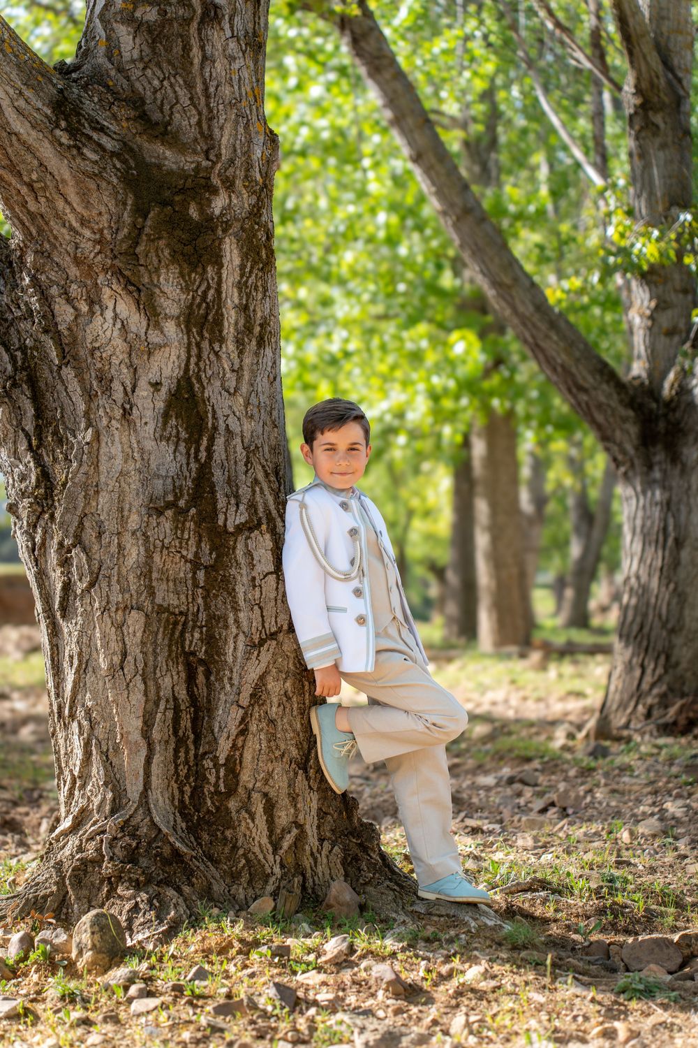A young boy in a white formal suit leans against a large tree trunk in a lush green park setting.