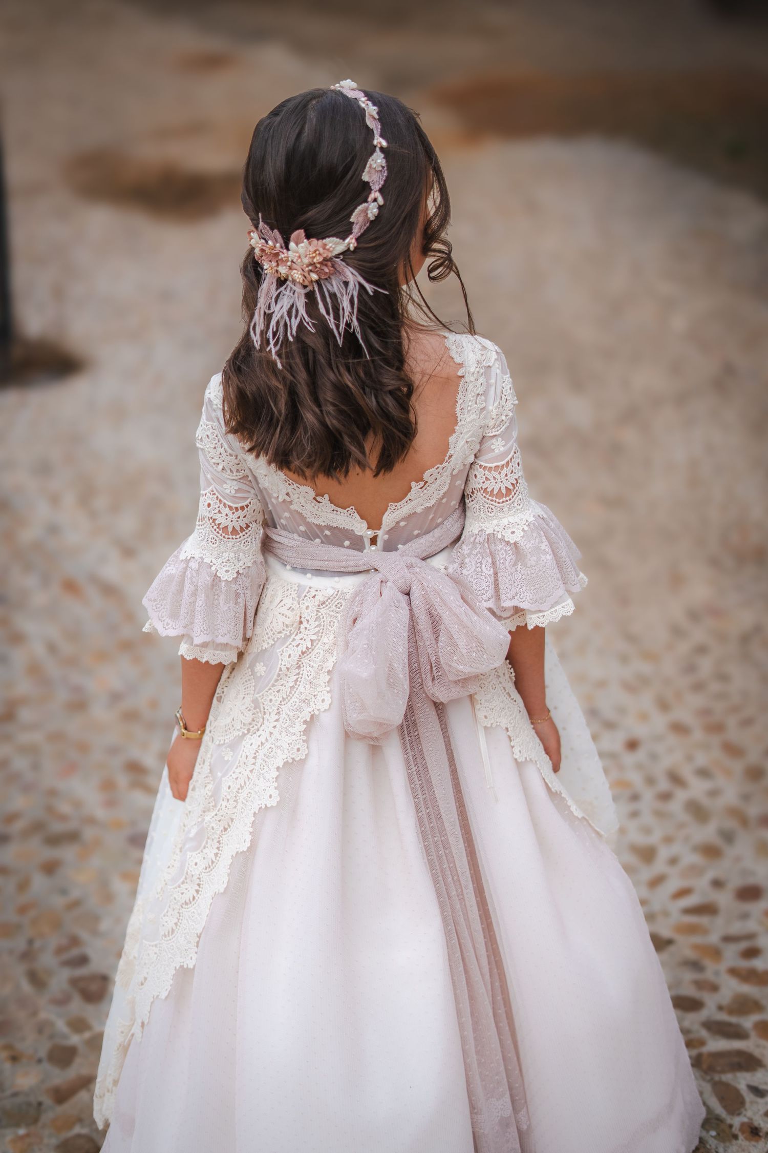Young girl in elegant white lace dress with pink bow and floral headpiece, viewed from behind on cobblestone path.