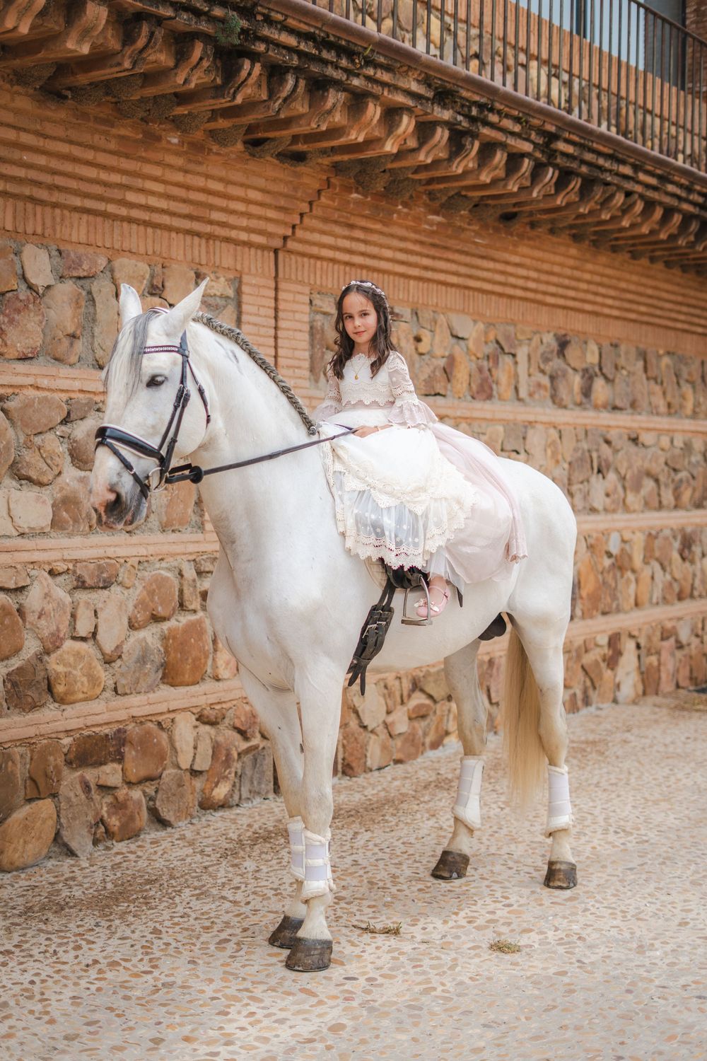 Young girl in white dress riding a white horse beside a stone wall in an elegant equestrian setting.