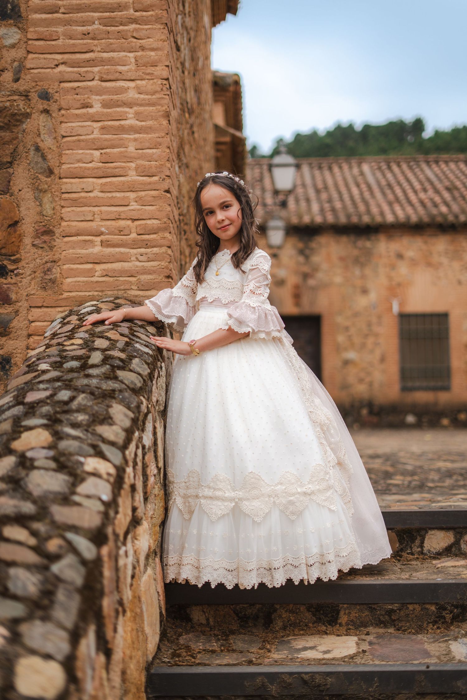 Young girl in white lace communion dress poses on stone steps beside rustic brick building in historic village.