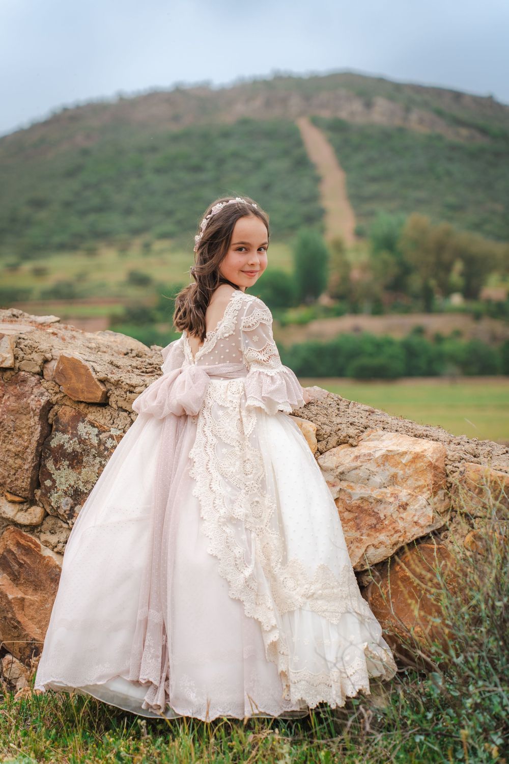 Young girl in elegant white lace ball gown poses outdoors near stone wall with green hills in background.