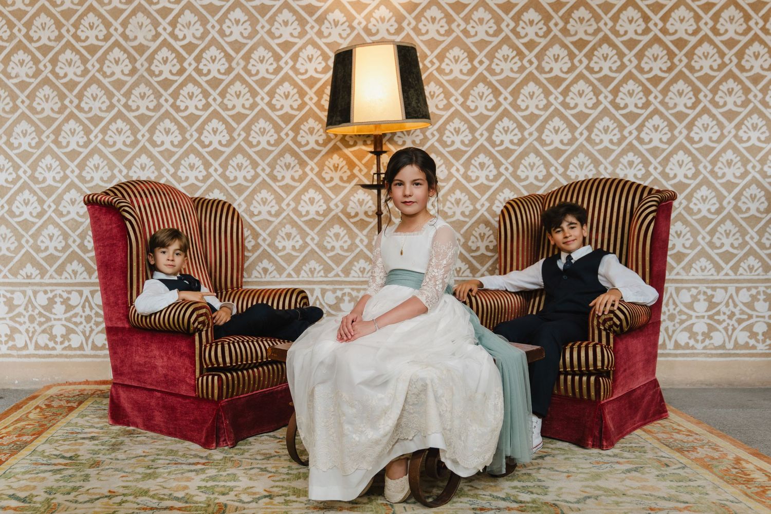 Three children in formal attire pose elegantly in ornate velvet armchairs against a vintage floral wallpaper backdrop.