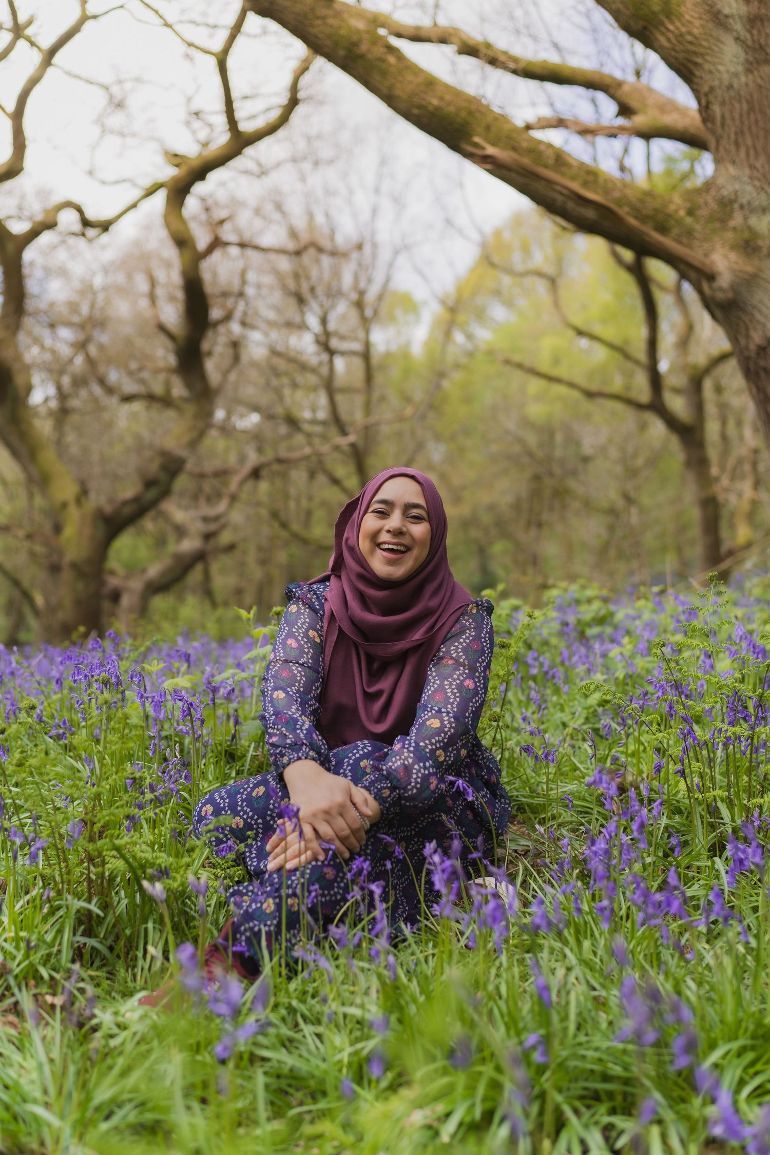 Someone wearing a purple floral dress and burgundy hijab sits among vibrant bluebells in a spring woodland setting.