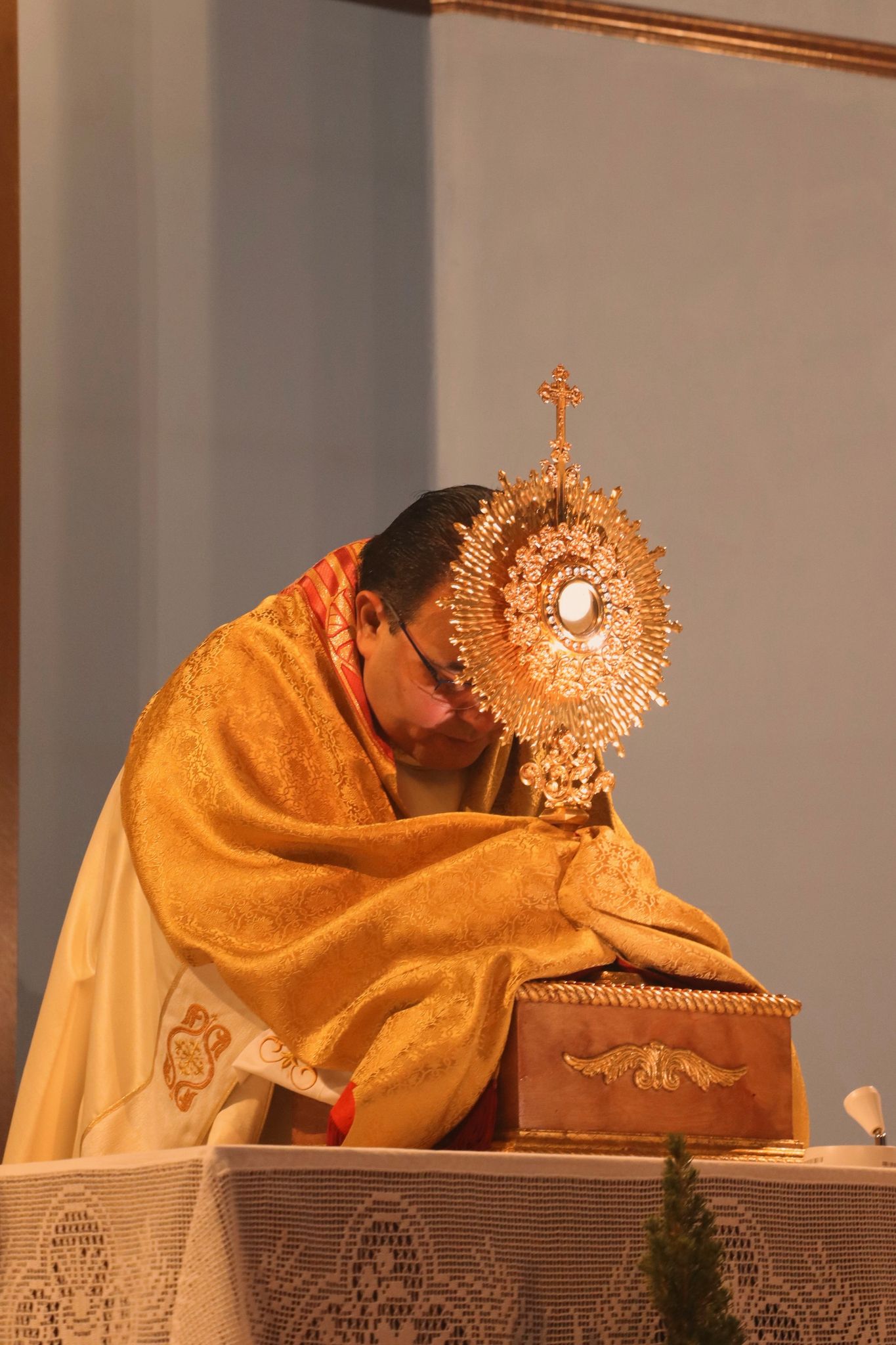A Catholic priest in gold vestments bows before a golden monstrance during Eucharistic adoration in a church service.