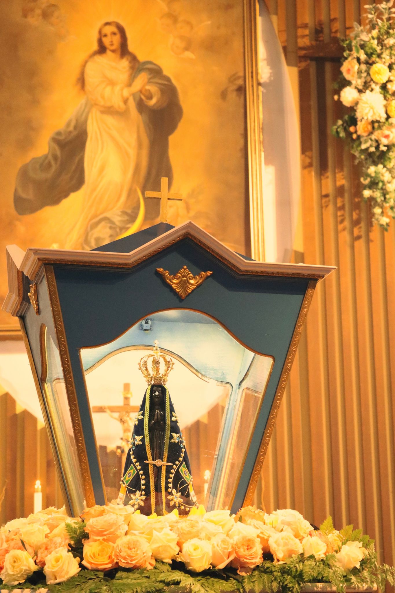 Religious altar with ornate lantern display holding statue of Virgin Mary, decorated with yellow roses and illuminated painting above.