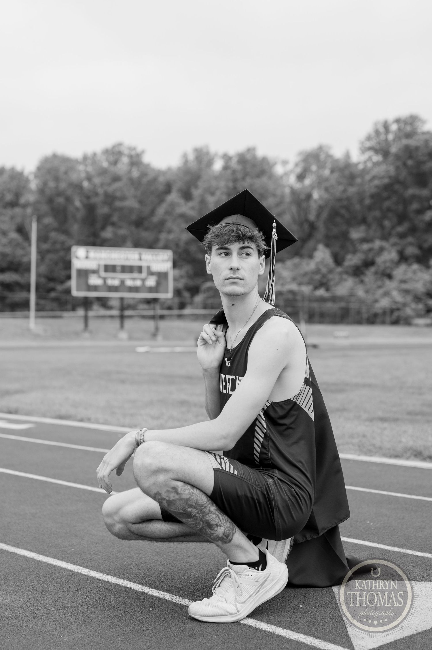 A black and white photo shows a graduate posing on a track field wearing academic regalia.