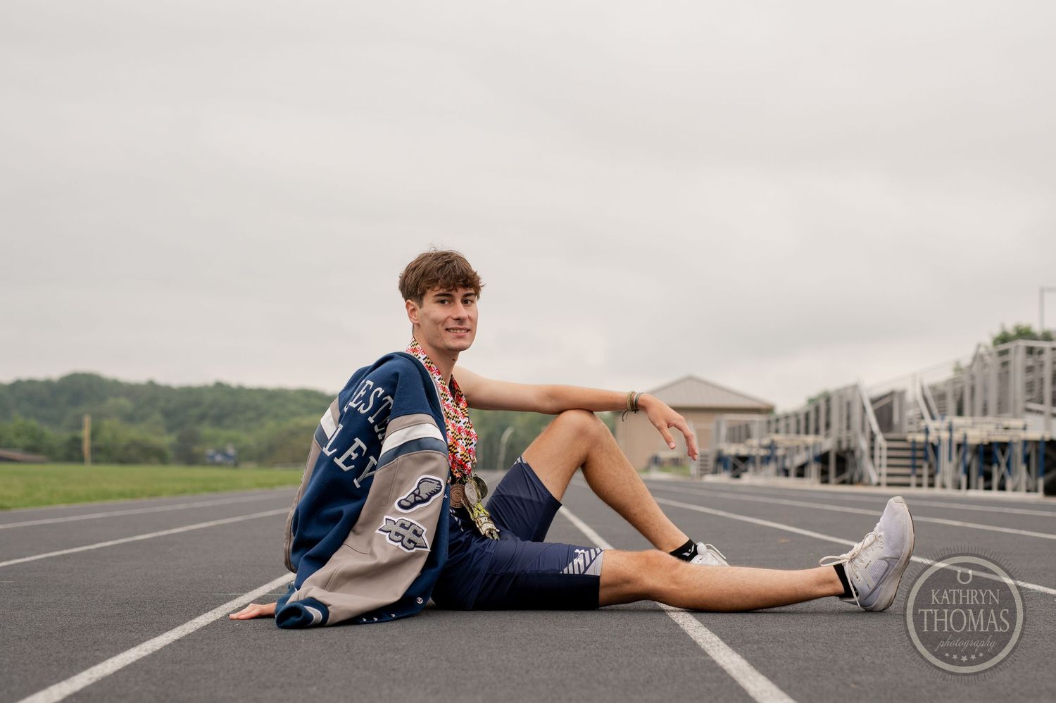 Athlete in navy uniform sits casually on running track with bleachers in background.