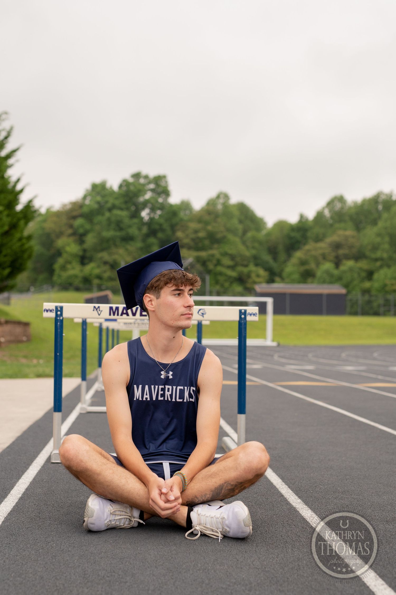 Graduate in navy uniform sits cross-legged on track near hurdles.