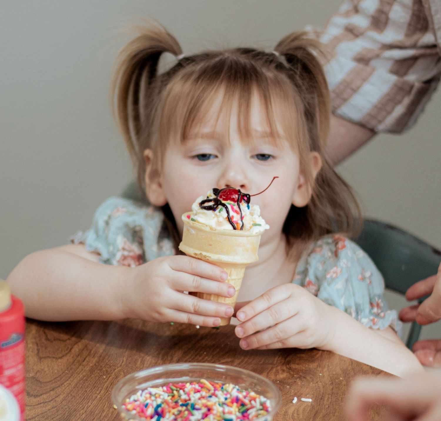 Child enjoying a vanilla ice cream cone topped with chocolate syrup and sprinkles while seated at a wooden table.