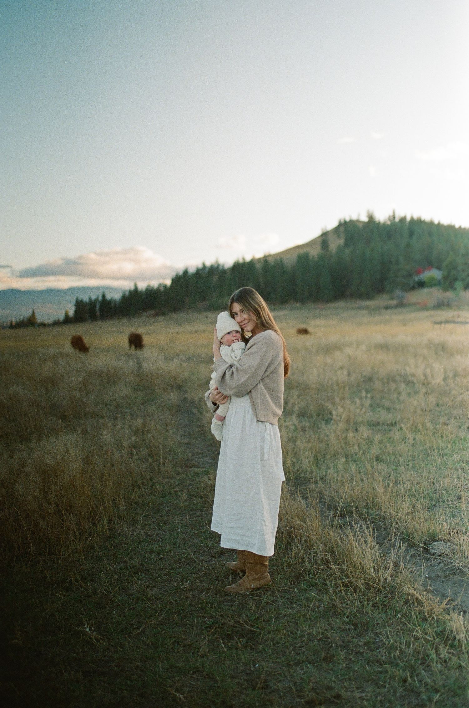 A serene mountainside scene with someone in a white dress and sweater standing in golden grassland at sunset.