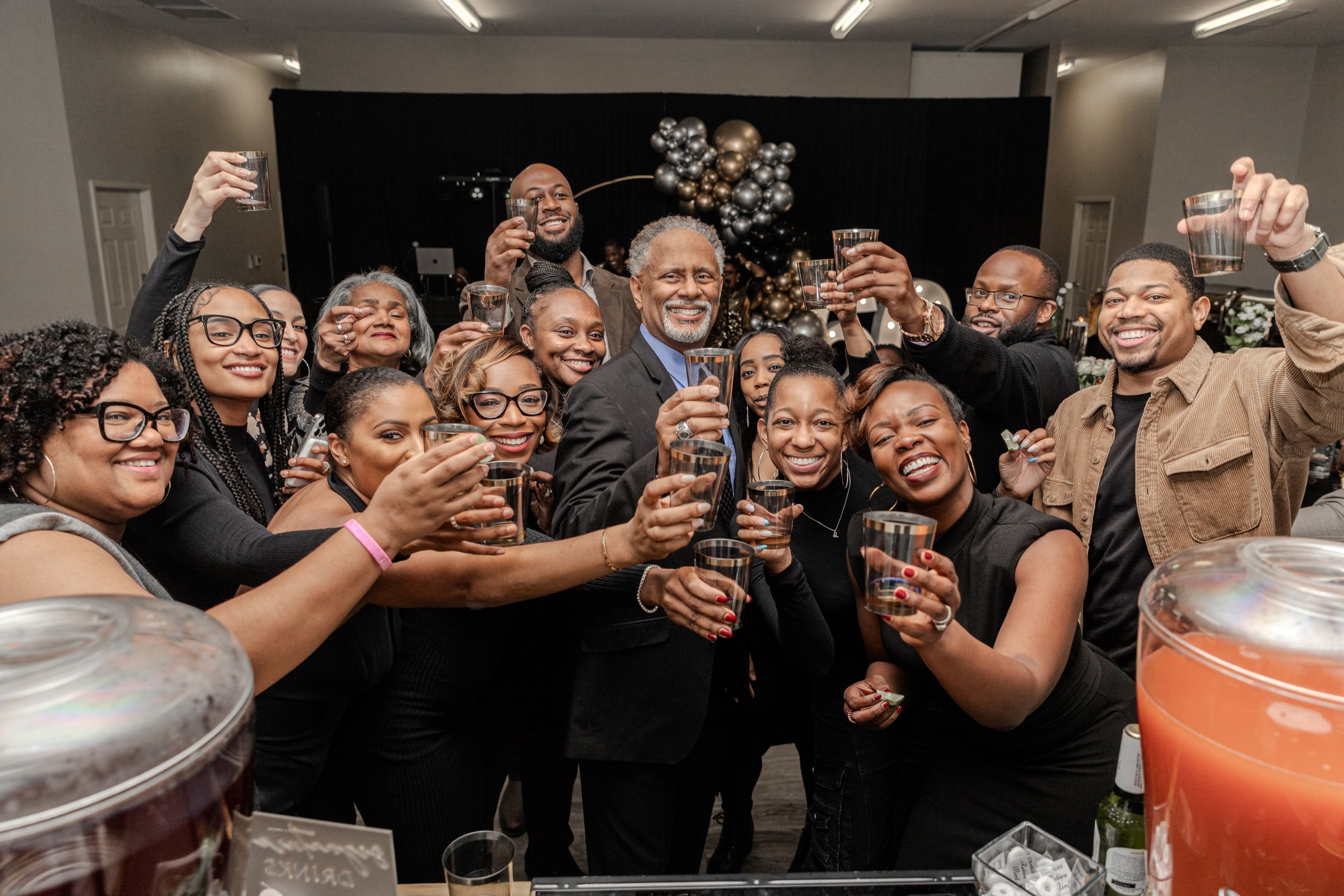 Group toasting with drinks at celebration event against black backdrop.