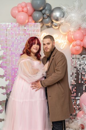 Couple in elegant attire posing under pink and silver balloon arch at celebration.
