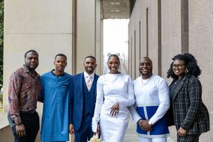 Wedding party in formal attire standing together against white building exterior.