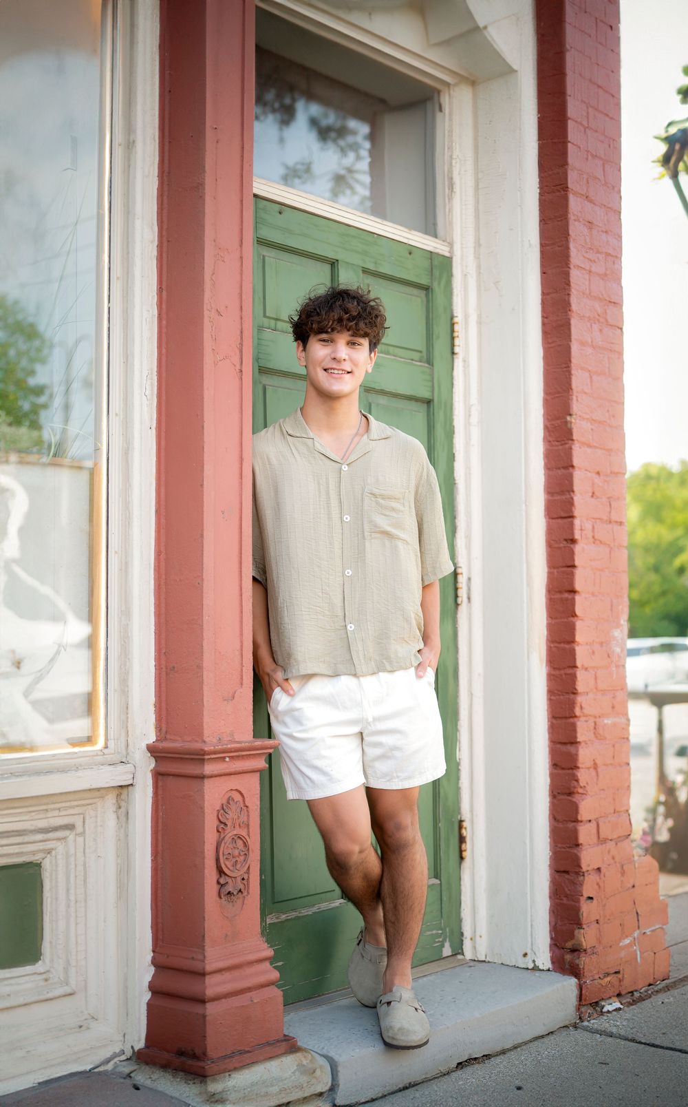 Someone poses in a beige shirt and white shorts against a green door with pink brick columns.