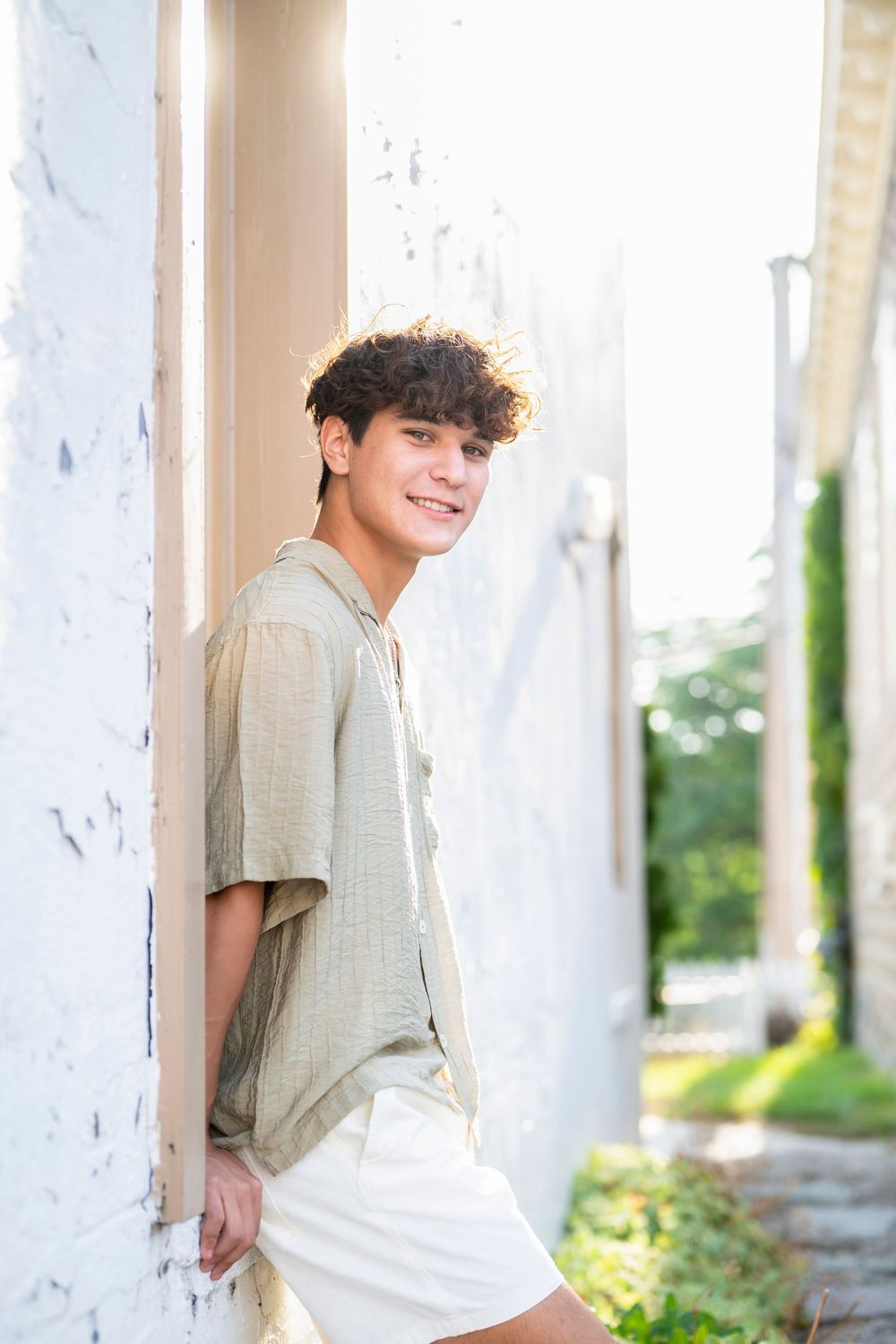 Someone leans against a white wall wearing a beige shirt and white shorts in summer lighting.
