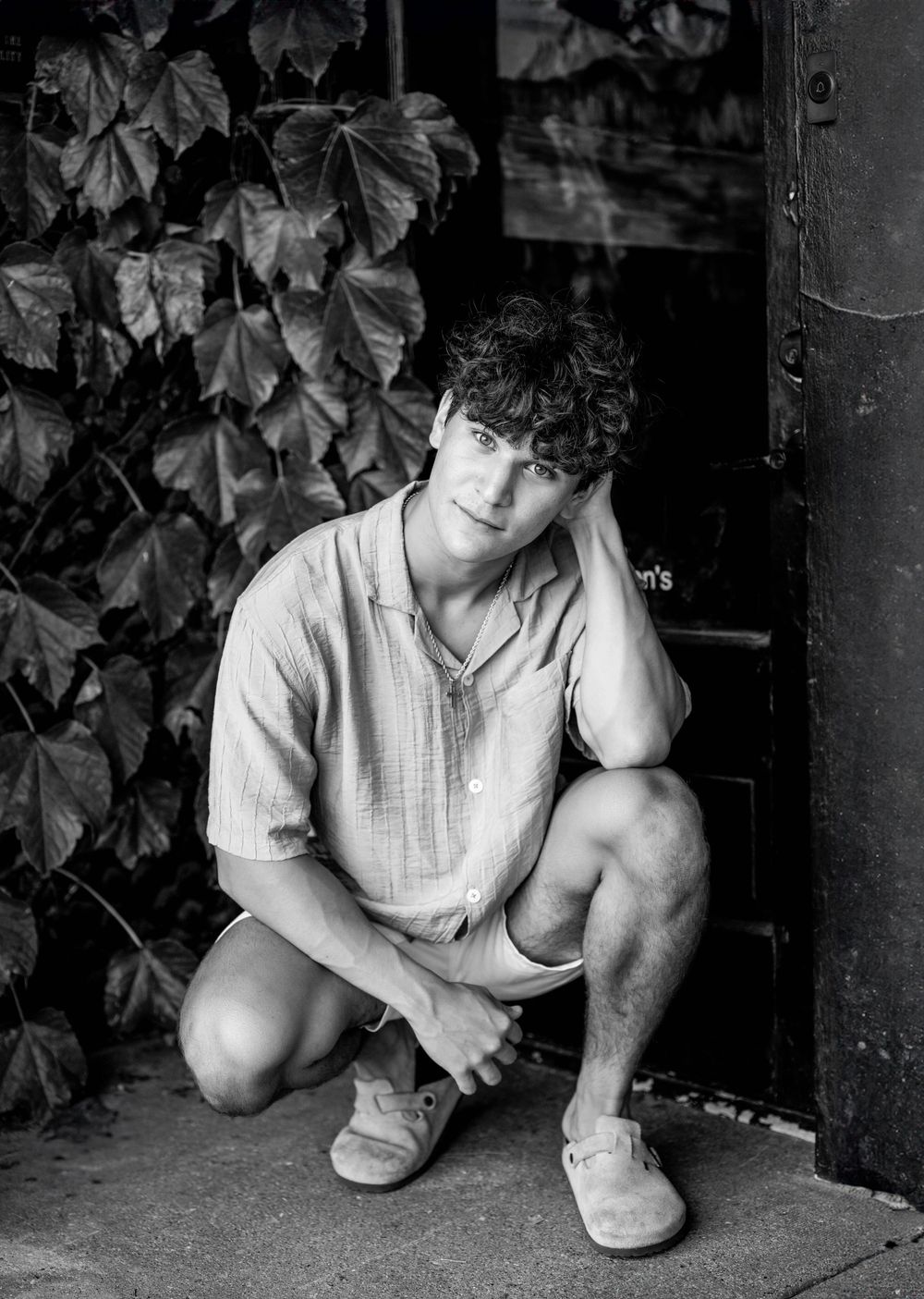 A black and white portrait of someone crouching near ivy-covered walls.