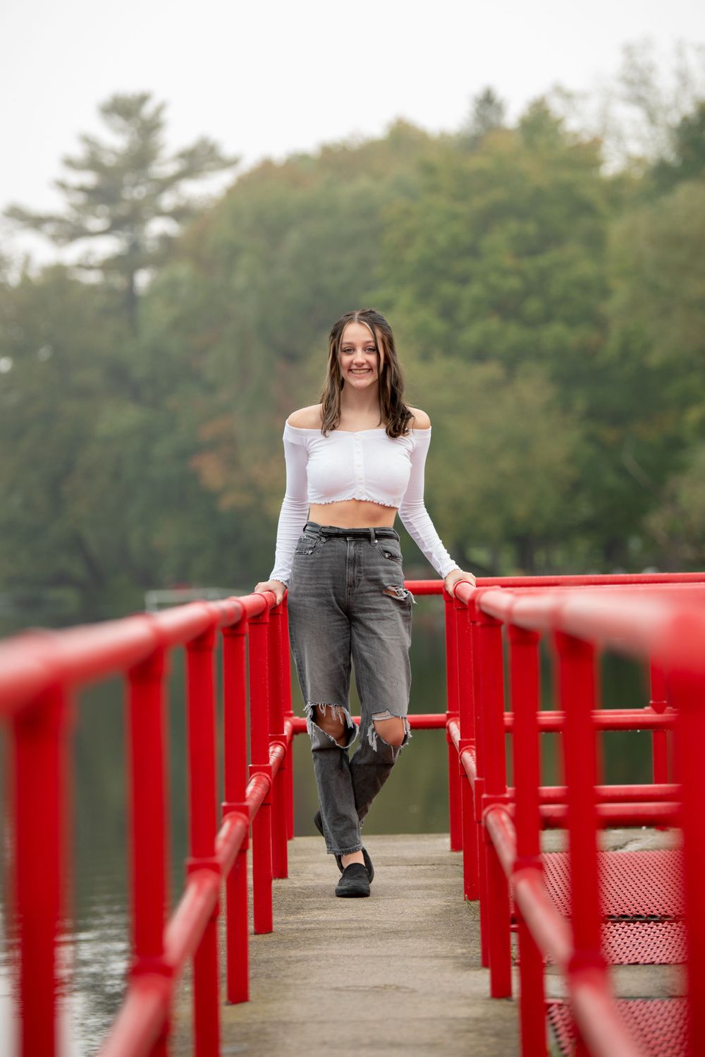 Person in white top and gray pants poses on red bridge railing over water.