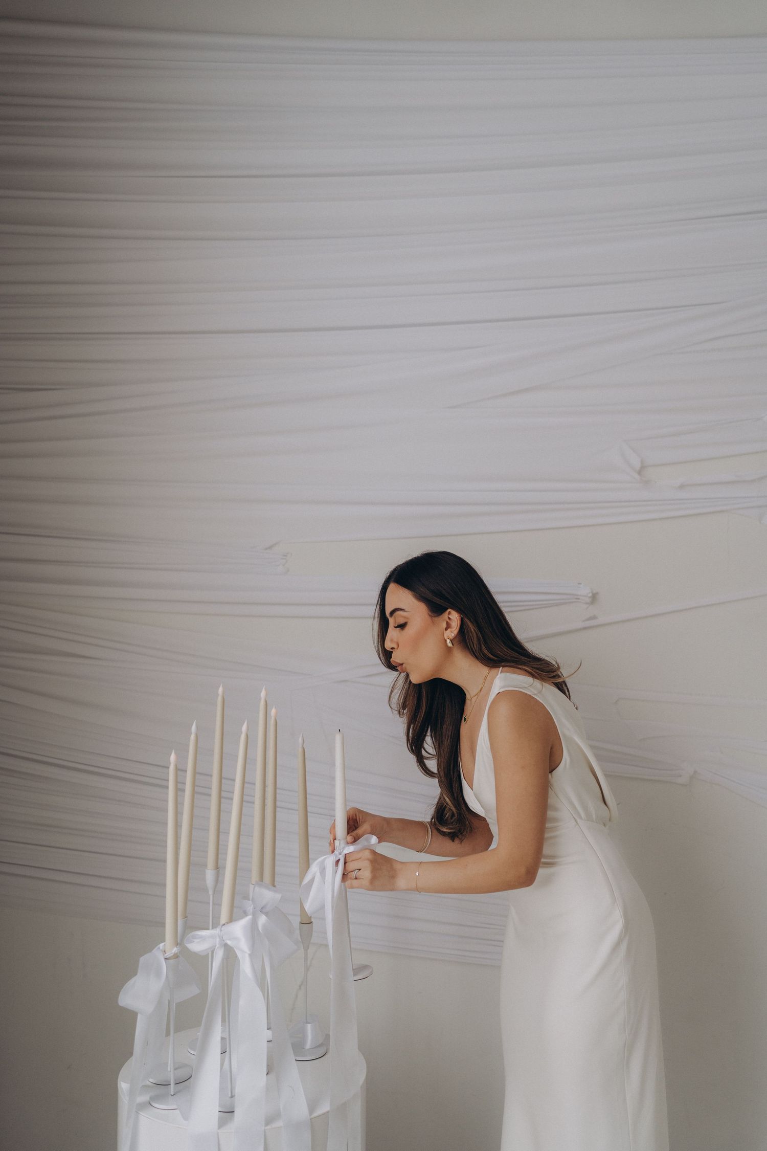 A woman in a flowing white dress lights elegant white candles during an ethereal indoor photoshoot.