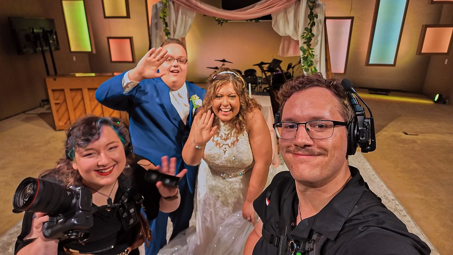 Group of people pose together on a TV studio set with wood panels and colorful wall decor in the background.