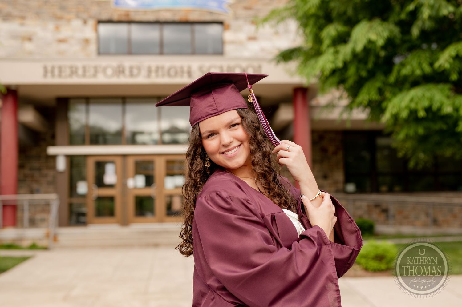Graduate in burgundy cap and gown smiles outside Hereford High School entrance.