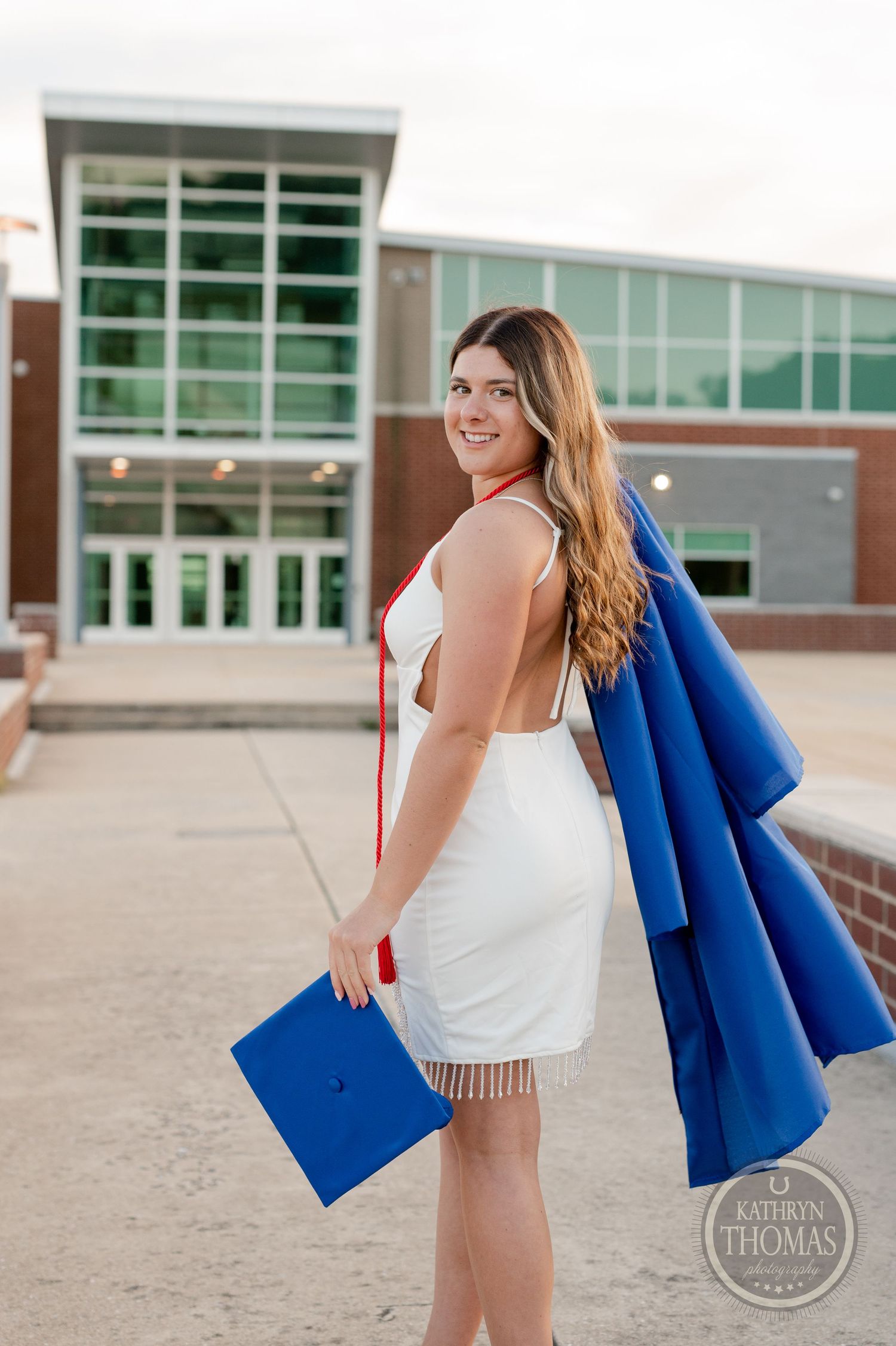Graduate in white dress carries blue graduation gown and cap while standing outside school building.