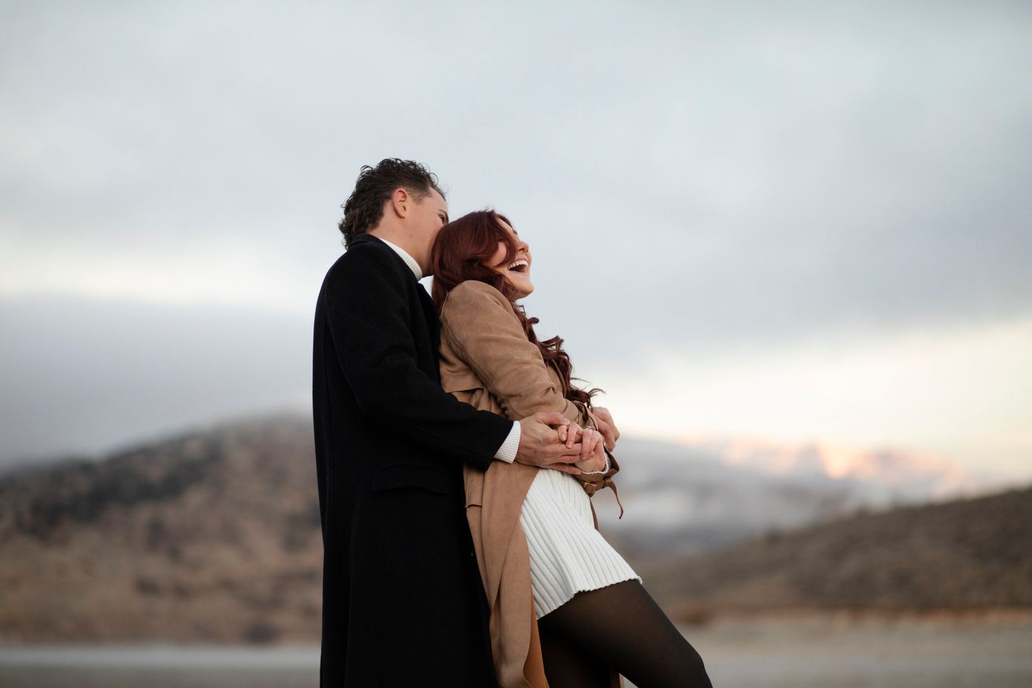 He runs up and grabs her from behind. He lifts her and she laughs.