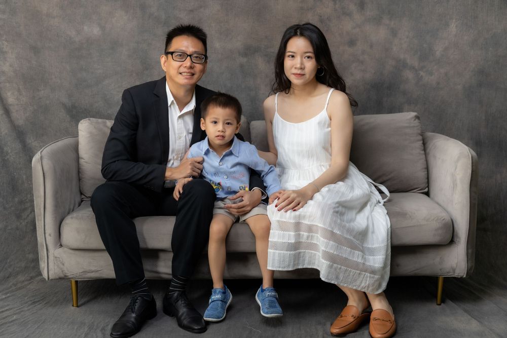A family of three sits together on a modern grey couch wearing formal and casual attire.
