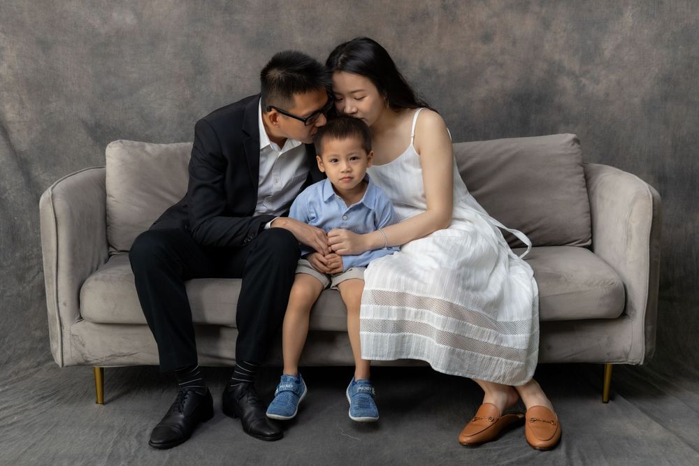 An intimate family portrait showing parents and child sharing a moment on a grey sofa.