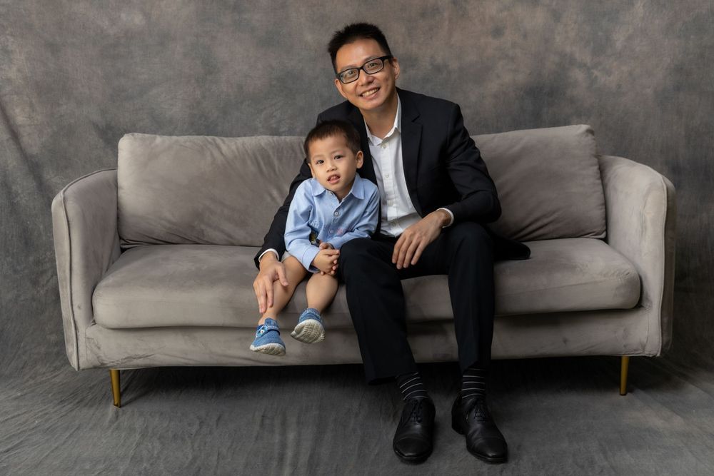 A parent and small child sit together on a gray couch against a textured dark background.