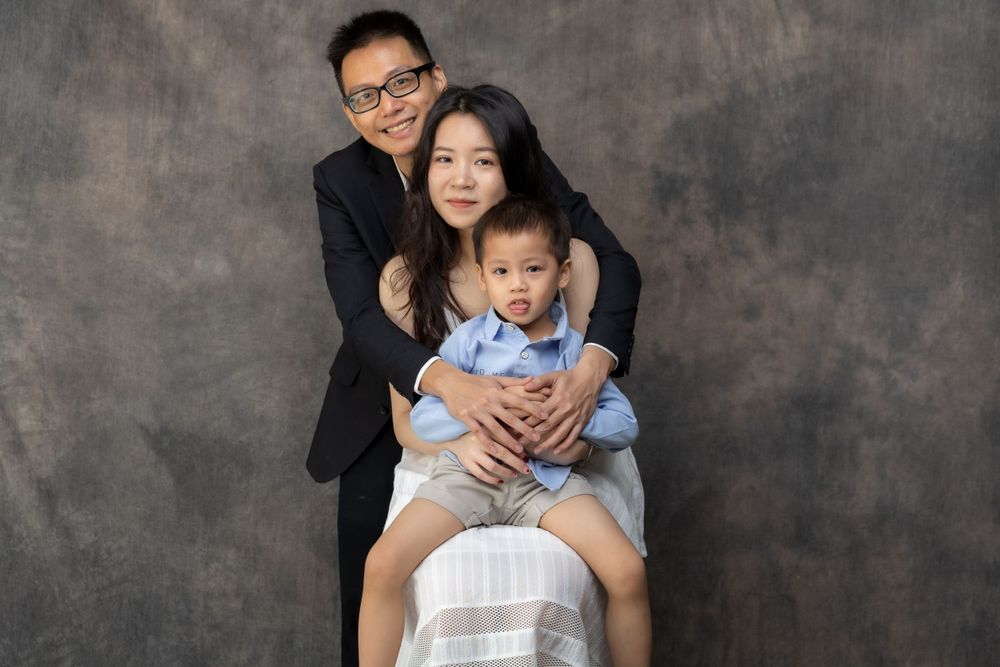 A family of three poses together against a mottled gray background in a studio setting.