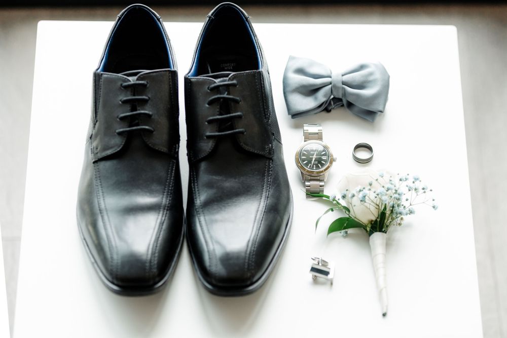 Black formal dress shoes displayed with a gray bow tie, rings, and boutonniere.