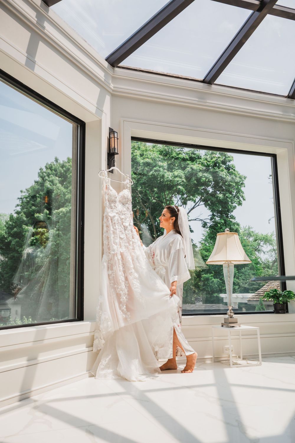 A bride in white robe inspects her lace wedding gown in a bright sunroom with floor-to-ceiling windows.