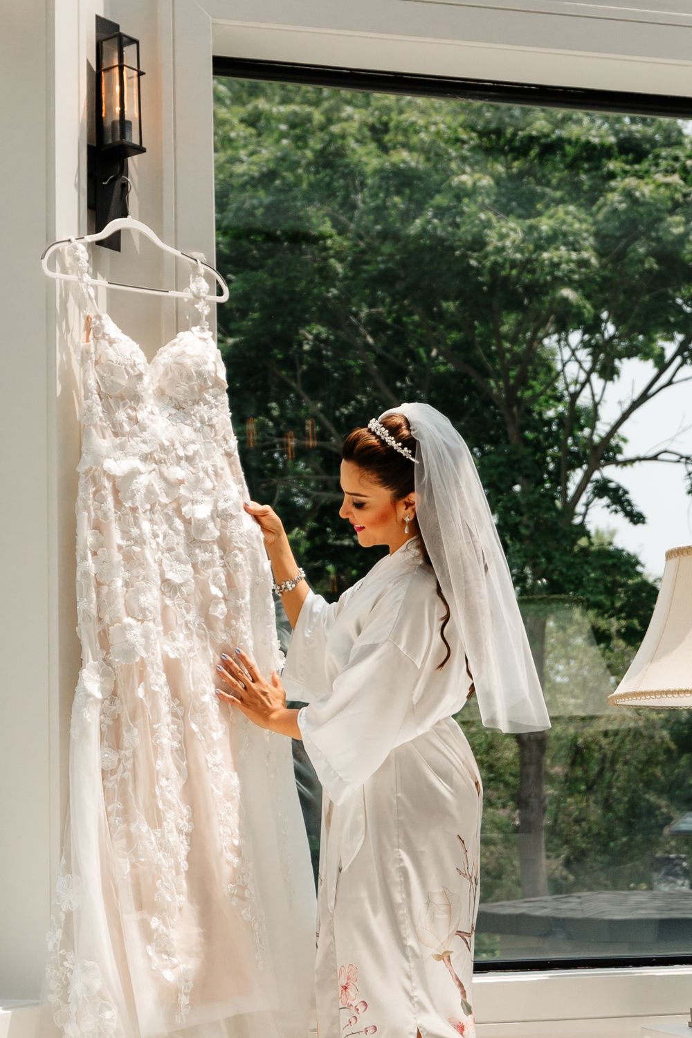 Bride in silk robe and veil touches her hanging lace wedding dress during morning preparations.