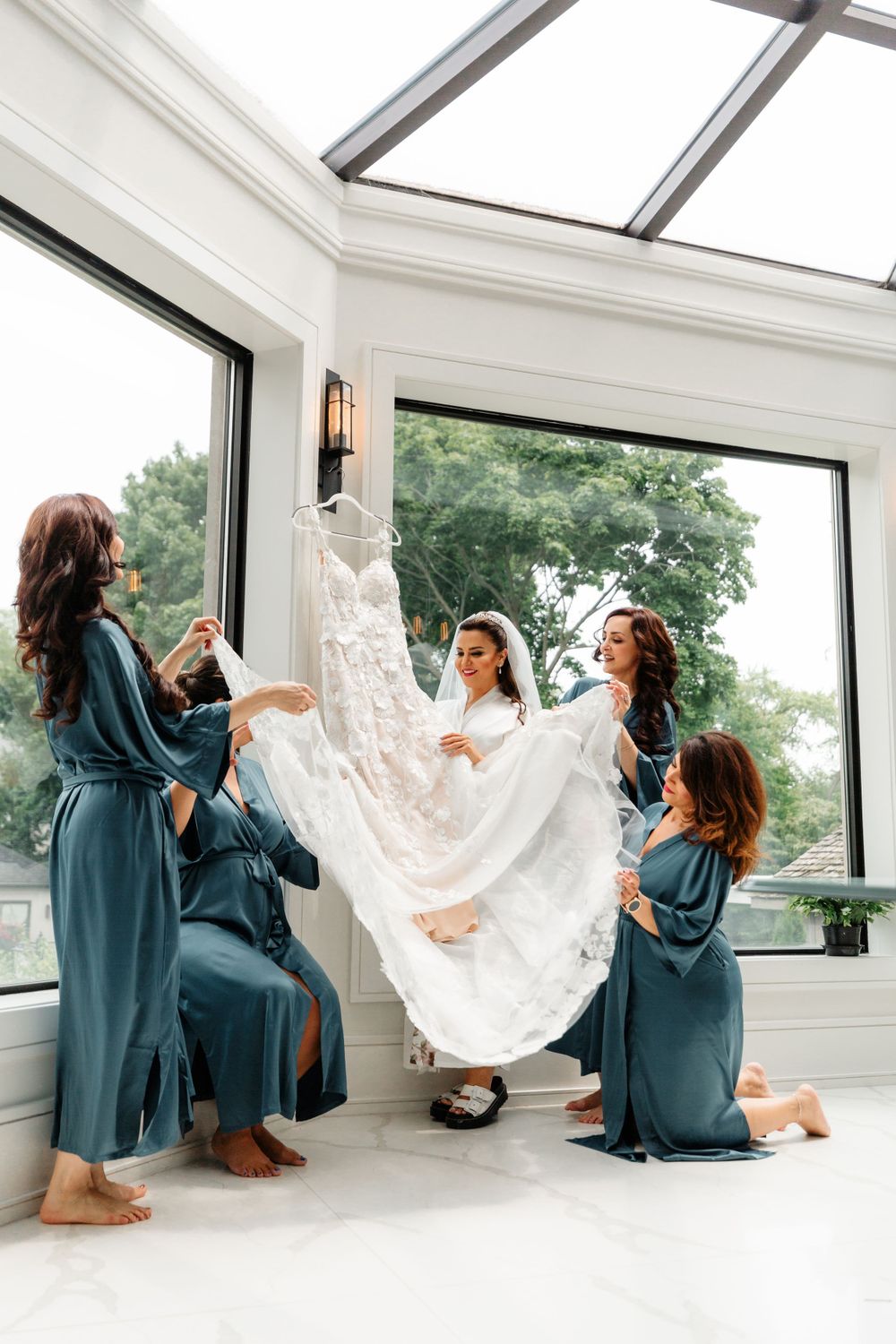 Bridesmaids in teal robes playfully holding up a lace wedding dress in a bright sunlit room with large windows.