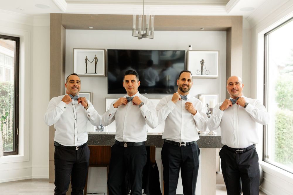 Four groomsmen in coordinated attire adjusting their bow ties for a fun photo.