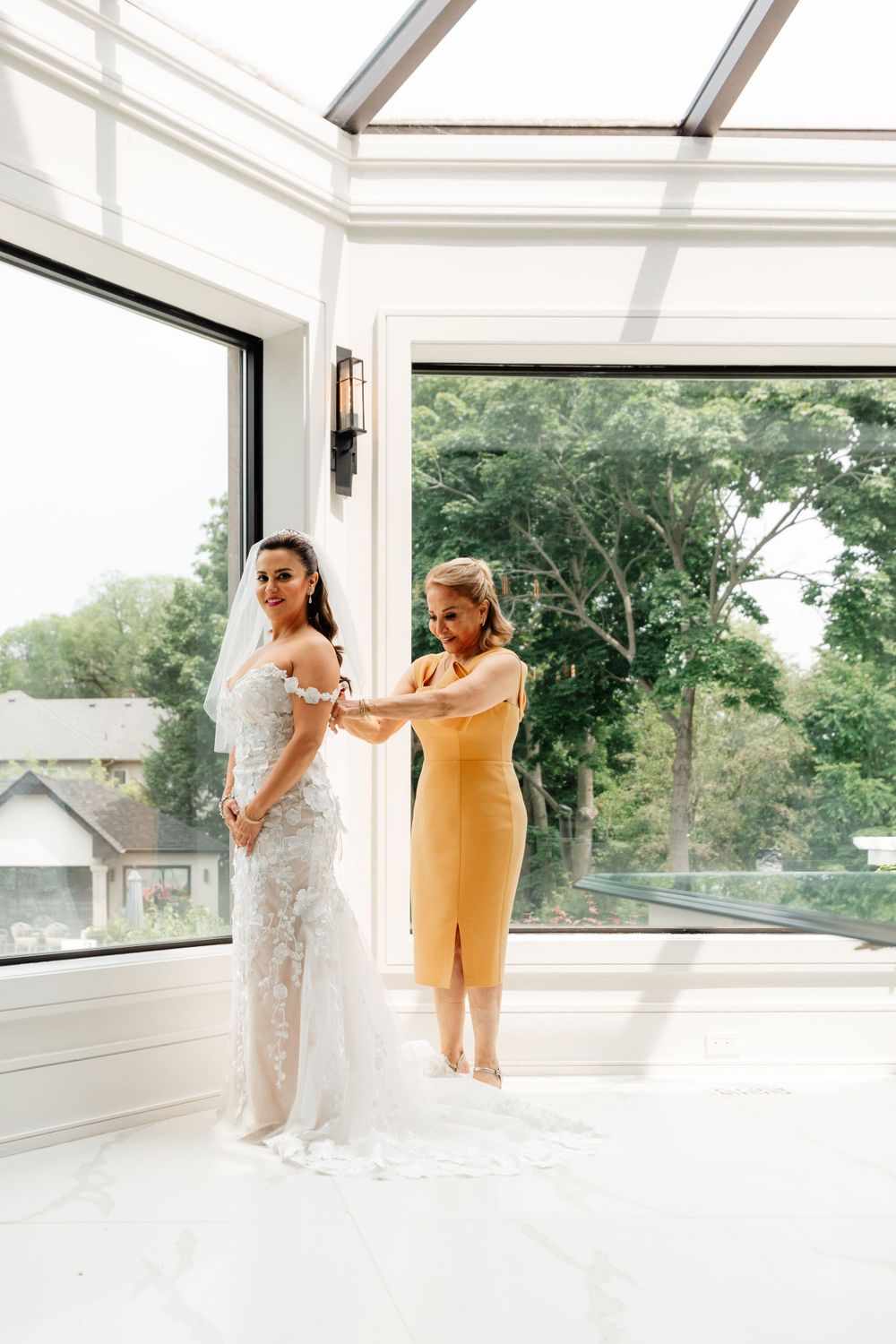 A wedding dress fitting in a white room with glass windows and green foliage visible outside.