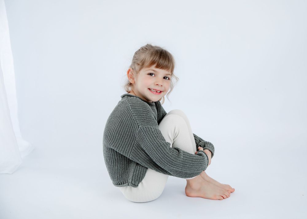 Child in cozy gray sweater sitting on white floor in studio setting.