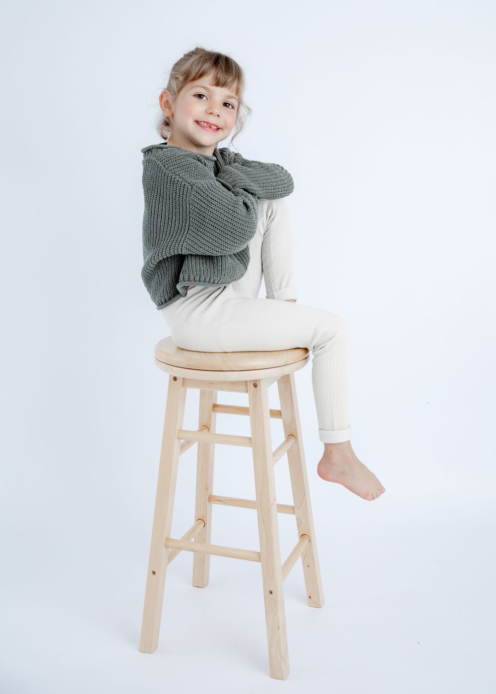 Child in gray sweater and white pants sitting casually on wooden stool against white background.