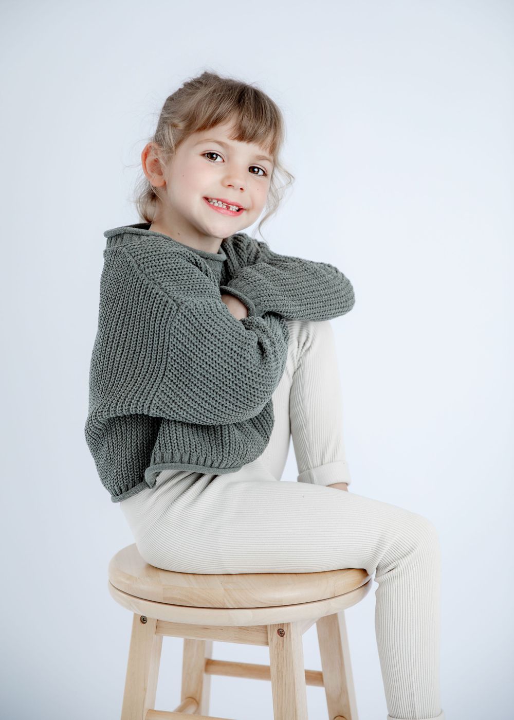 Casual pose on wooden stool wearing gray knit sweater in bright studio setting.